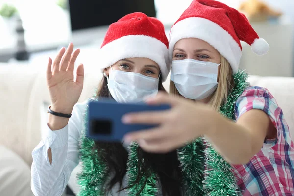 Dos mujeres jóvenes con sombreros de santa y máscaras protectoras en las caras están sosteniendo el teléfono en el retrato de casa — Foto de Stock