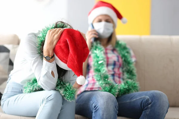 Kvinna i Santa claus hatt håller huvudet med händerna medan vän i skyddsmask talar i telefon — Stockfoto