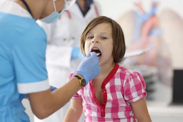 Dokter berjas biru melihat tenggorokan anak dengan tongkat kayu. — Stok Foto