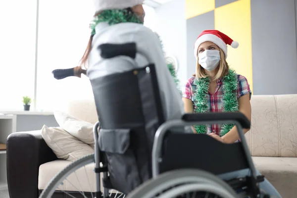 Жінка в капелюсі Санта Клауса і з мішурою навколо шиї в захисній масці на її обличчі спілкується зі своїм другом у інвалідному кріслі — стокове фото