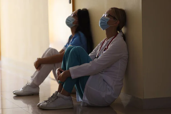 Joven doctora y enfermera en máscaras médicas se sientan en el pasillo del hospital — Foto de Stock