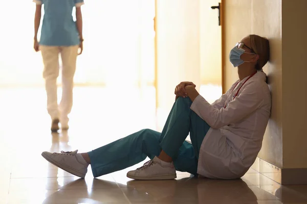 Lékař v lékařské masce sedí na podlaze v nemocniční chodbě a chůzi zdravotní sestra v pozadí — Stock fotografie