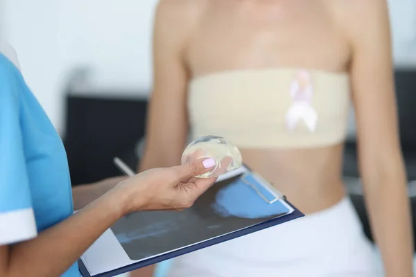 Doctors hands are holding breast implant in front of clipboard with patient in background — Stock Photo, Image