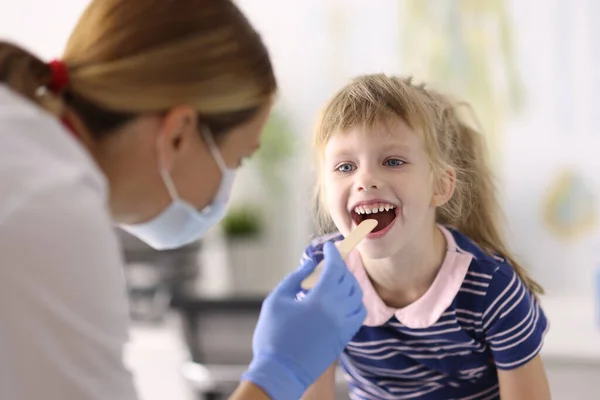 Medico pediatra donna in maschera protettiva medica e guanti di gomma esamina la gola della bambina spatola di legno ritratto — Foto Stock