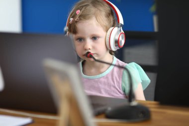 Küçük kız kulaklığı ve mikrofonuyla bilgisayarın başında oturuyor.