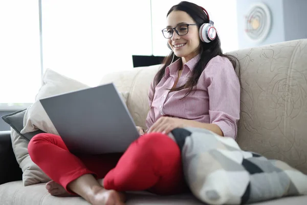 Woman in headphones with laptop on her lap is sitting on couch and smiling.