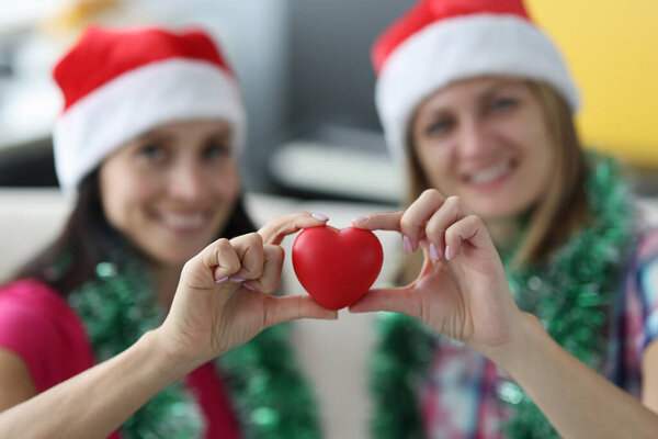 Two smiling woman in santa hats hold heart.