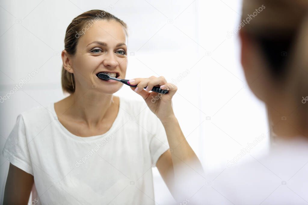 Woman in awhite T-shirt is brushing her teeth in front of mirror