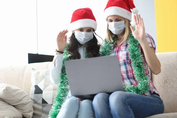 Duas mulheres em máscara protetora e chapéu de Papai Noel sentar lado a lado e acenar Olá. — Fotografia de Stock