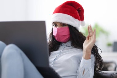 Noel Baba şapkalı kadın koruyucu maske takıyor gülümsüyor ve dizüstü bilgisayara el sallıyor..