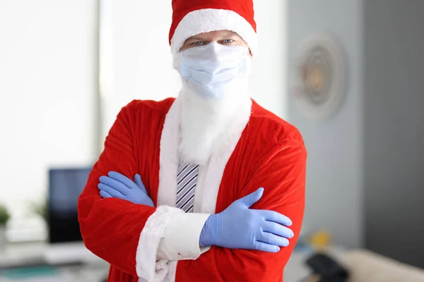 Homem em traje de Papai Noel e máscara médica protetora fica com braços dobrados. — Fotografia de Stock