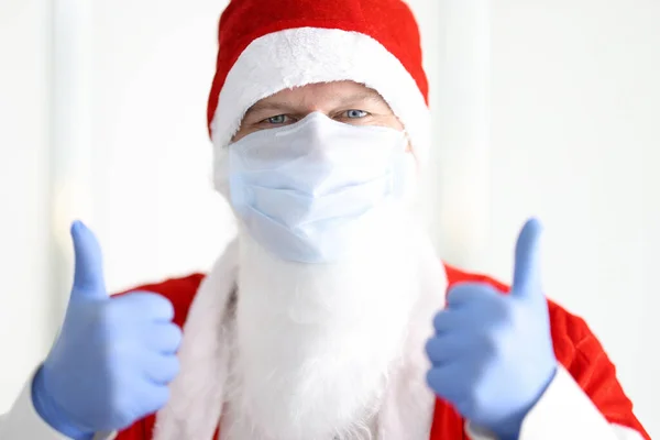 Homme en costume de Père Noël claus et masque médical de protection et des gants tient ses pouces vers le haut. — Photo