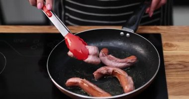 Kadın sosisleri fırının tepesindeki tavaya atmak için maşa kullanıyor. Hızlı yemek hazırlama süreci, tüketim için et kızartma eylemini vurguluyor