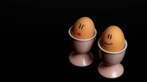 Emotional two boiled eggs in pink egg cups with drawn red lips and mustache isolated on black background