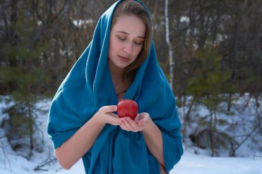 Ellerinde kırmızı elmayla mavi başlıklı bir kız. Kız ellerindeki elmaya bakıyor. Noel 'de kış ormanında bir kadın.