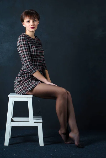 Una chica con el pelo corto en un vestido está sentado en una silla sobre un fondo oscuro —  Fotos de Stock