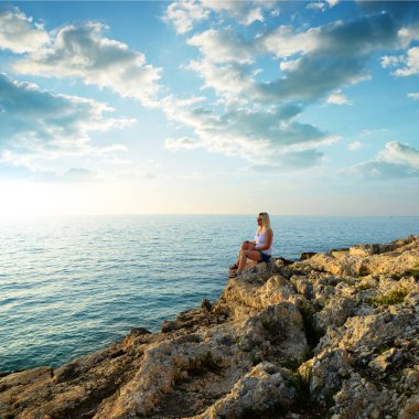 Bir uçurumun üzerinde oturan bir kadın denizin üzerinde gün batımını izlemek.