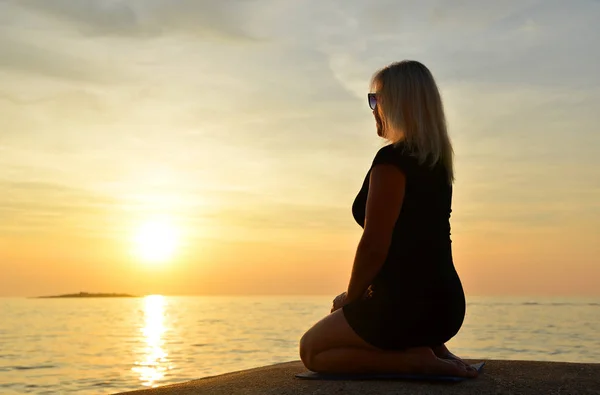 Woman sitting in yoga pose by the sea at sunset.