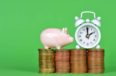The small pink piggy bank with pile of coins and white alarm clock on green background. Time to save money concept. clipart