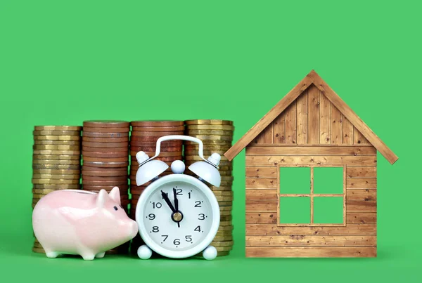 緑の背景に小さなピンクの貯金箱 コインの山 白い目覚まし時計と木製の家 新しい住宅にお金を節約するための時間 — ストック写真