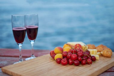 İki kadeh kırmızı şarap, açık havada servis edilen meyveler ve güzel mavi okyanus manzaralı..