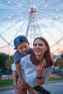 Neşeli oğul, parkta dönme dolabın arkasında yürürken annesine sarılıyor..