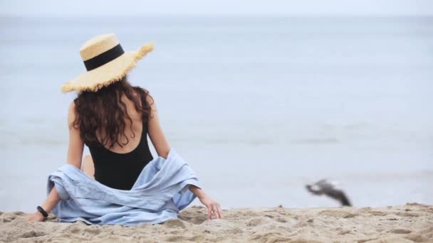 Chica en un sombrero se sienta en la playa — Vídeos de Stock