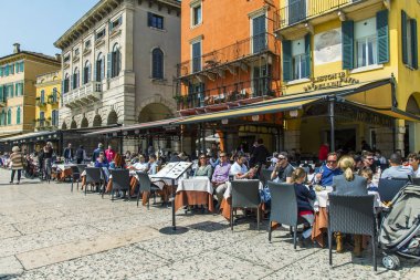 Verona, İtalya, 27 Nisan 2019 tarihinde. İnsanlar şehrin merkezi meydanlarından biri olan Piazza Bra Meydanı'ndaki sokak kafesinin masalarında yemek yiyip dinleniyor.