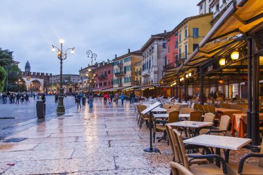 Verona, İtalya, 27 Nisan 2019 tarihinde. İnsanlar, akşam saatlerinde yağmurdan sonra şehrin merkezi meydanlarından biri olan Piazza Bra Meydanı'na doğru gidiyor. Bir tente altında pitoresk sokak kafe Tablolar.