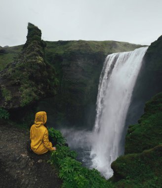 Woman in yellow raincoats looking on scenic high waterfall clipart