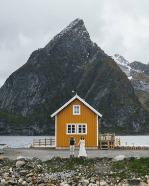 Sahilde kayalık Kvalvika turuncu ev ve dağ ile arka plan, Norveç üzerinde duran mutlu düğün çift yolcuları 