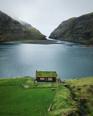Sisli yeşil dağlar arasında yeşil çatılı, ıssız deniz evi, Faroe adaları, Saksun köyü. Dikey doğa fotoğrafı.