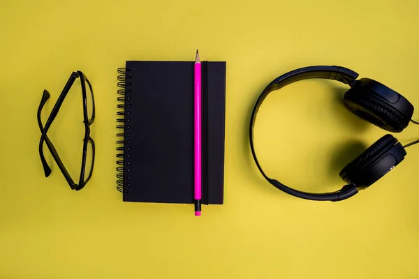 Music headphones, eyeglasses, notebook and pencil on bright yellow background