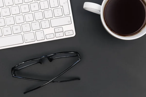 Minimal office desk computer keyboard coffee reading glasses on dark background