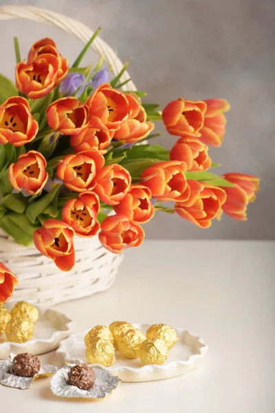 Candy in white dishes and a bouquet of orange tulips on light dyed wooden surface.