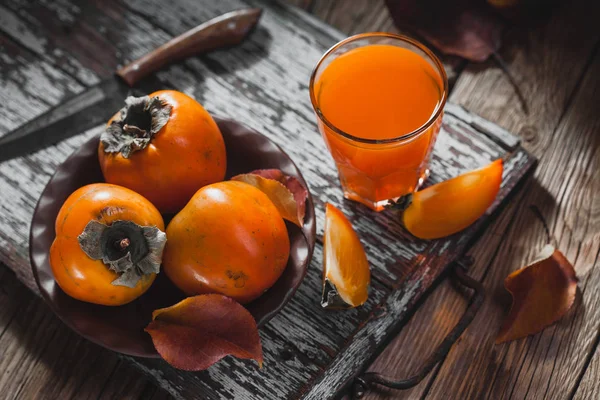 茶色の木製のテーブルに茶色のプレートでフレッシュ ジュースと熟したオレンジ色の柿と柿のガラスを残します 新鮮な果物 — ストック写真
