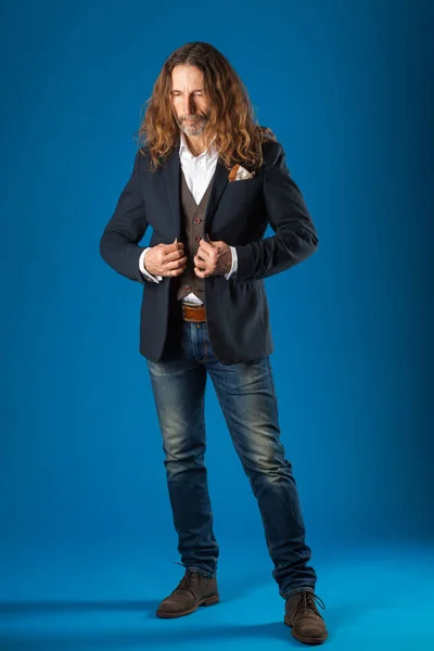 Uomo elegante dai capelli lunghi in jeans e giacca su uno sfondo blu . — Foto Stock
