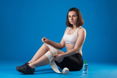 Esmer atlet kız mavi bir arka plan üzerinde bir bandaj ile bacak sarar.