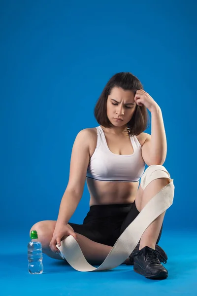 Brunette athlète fille enveloppe la jambe avec un bandage sur un fond bleu . — Photo