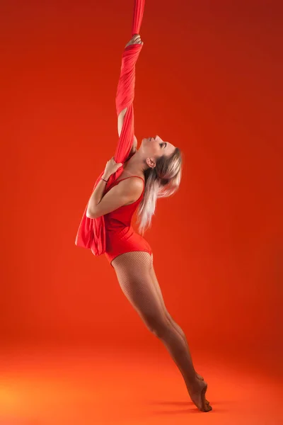 Uma jovem loira com cabelos longos em um terno vermelho realiza ginástica e exercícios em seda . — Fotografia de Stock