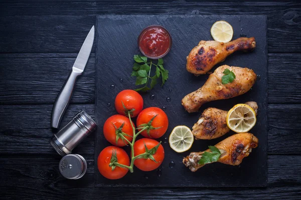 Cosce di pollo con pomodori, limone, prezzemolo e ketchup su una lavagna nera — Foto Stock