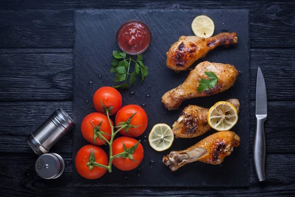Cosce di pollo con pomodori, limone, prezzemolo e ketchup su una lavagna nera — Foto Stock