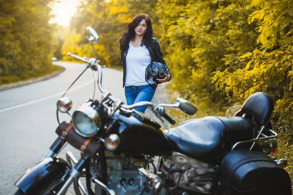 Attractive brunette motorcyclist standing near a motorcycle On the Sunset. — Stock Photo, Image