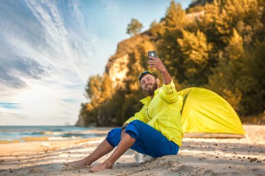 sakallı adam kumlu bir plajda bir çadır ile selfie yapar.