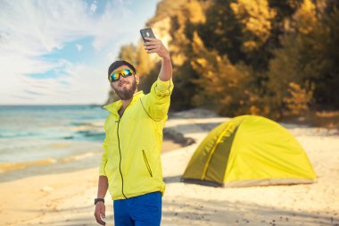 sakallı adam kumlu bir plajda bir çadır ile selfie yapar.