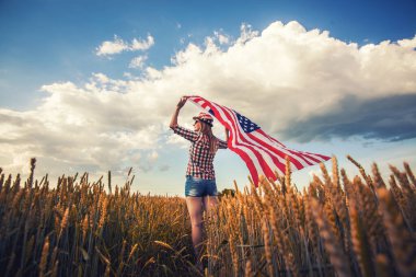 Güzel bir genç kız buğday tarlasında rüzgarda Amerikan bayrağını tutuyor. Mavi gökyüzüne karşı yaz manzarası. Bağımsızlık Günü 4 Temmuz