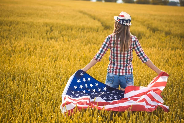 小麦の分野でアメリカの国旗を持っている美しい若い女の子 青空に対する夏の風景 独立記念日7月4日 — ストック写真