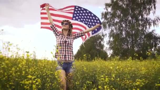 菜の花畑でアメリカ国旗を掲揚している美しい少女 青空に対する夏の風景 独立記念日7月4日 ロイヤリティフリーのストック動画