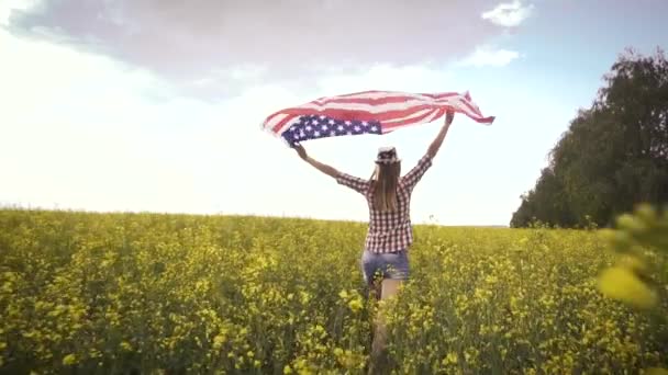 菜の花畑でアメリカ国旗を掲揚している美しい少女 青空に対する夏の風景 独立記念日7月4日 ストック動画