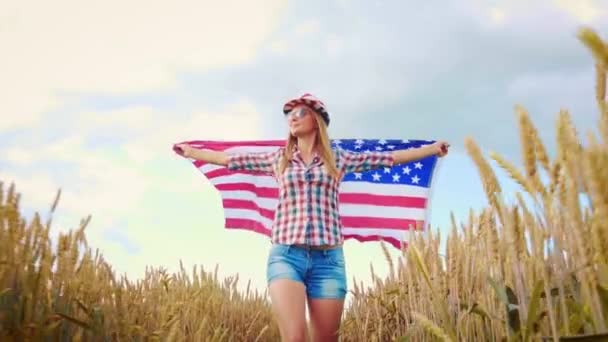 Bella ragazza che tiene una bandiera americana sul vento in un campo di grano. Paesaggio estivo contro il cielo blu. — Video Stock
